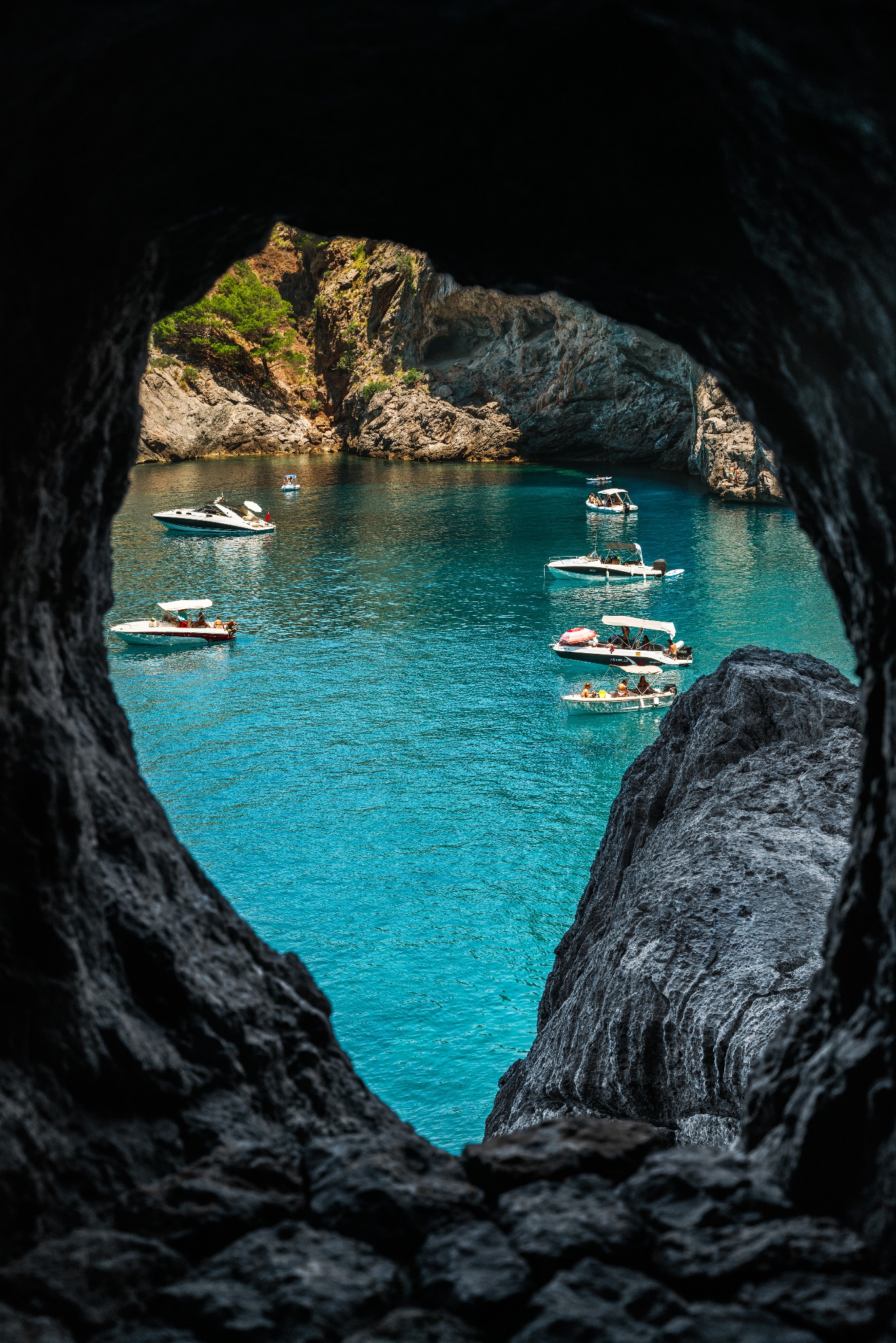 Widok z jaskini na turkusową zatokę z łodziami motorowymi i skalistym brzegiem porośniętym zielenią.