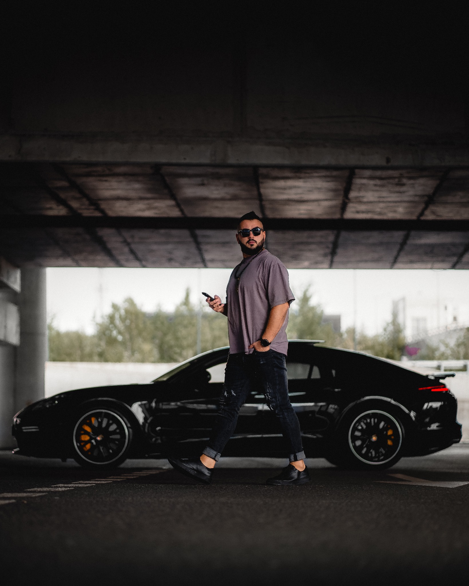 Mężczyzna w okularach przeciwsłonecznych idzie obok czarnego sportowego samochodu pod betonowym wiaduktem, trzymając telefon w ręku.