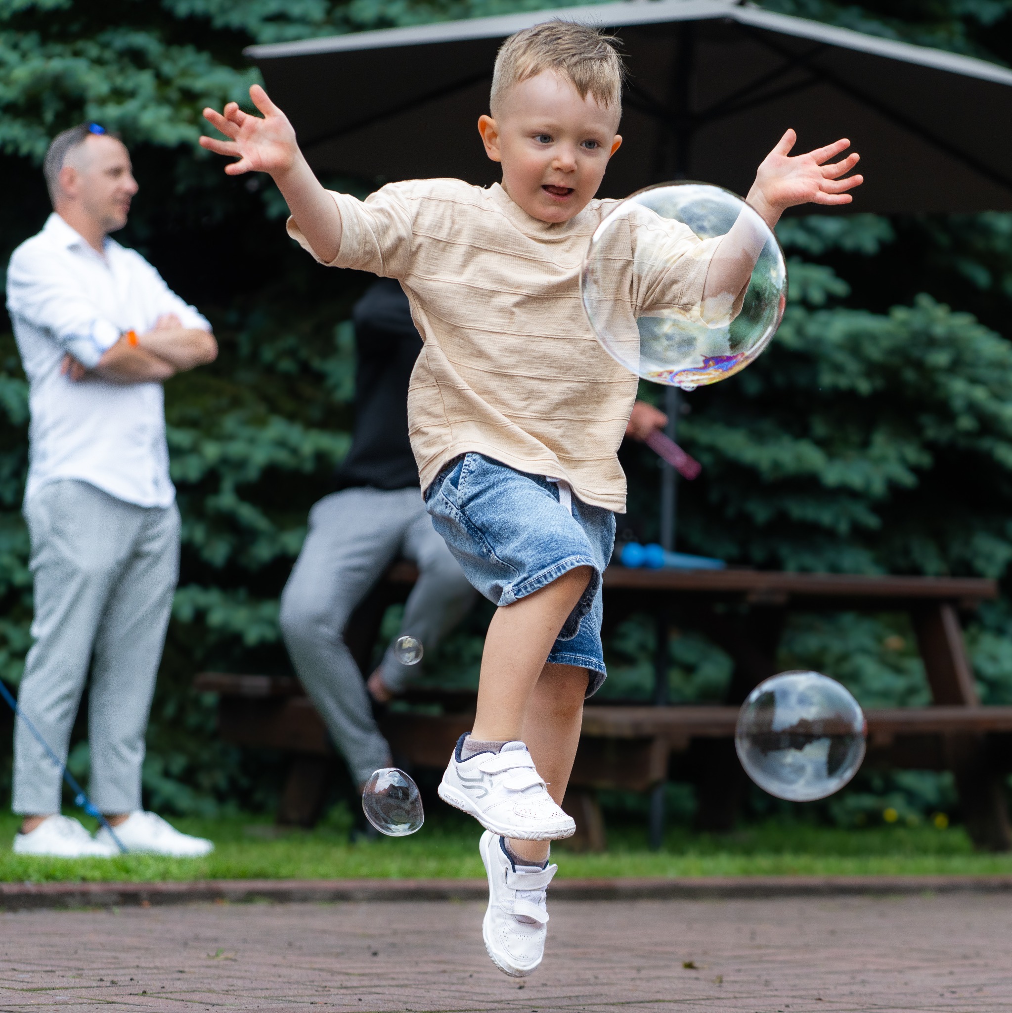 Chłopiec w beżowej koszulce i dżinsowych spodenkach skacze, łapiąc duże bańki mydlane, w tle obserwujący go mężczyźni.