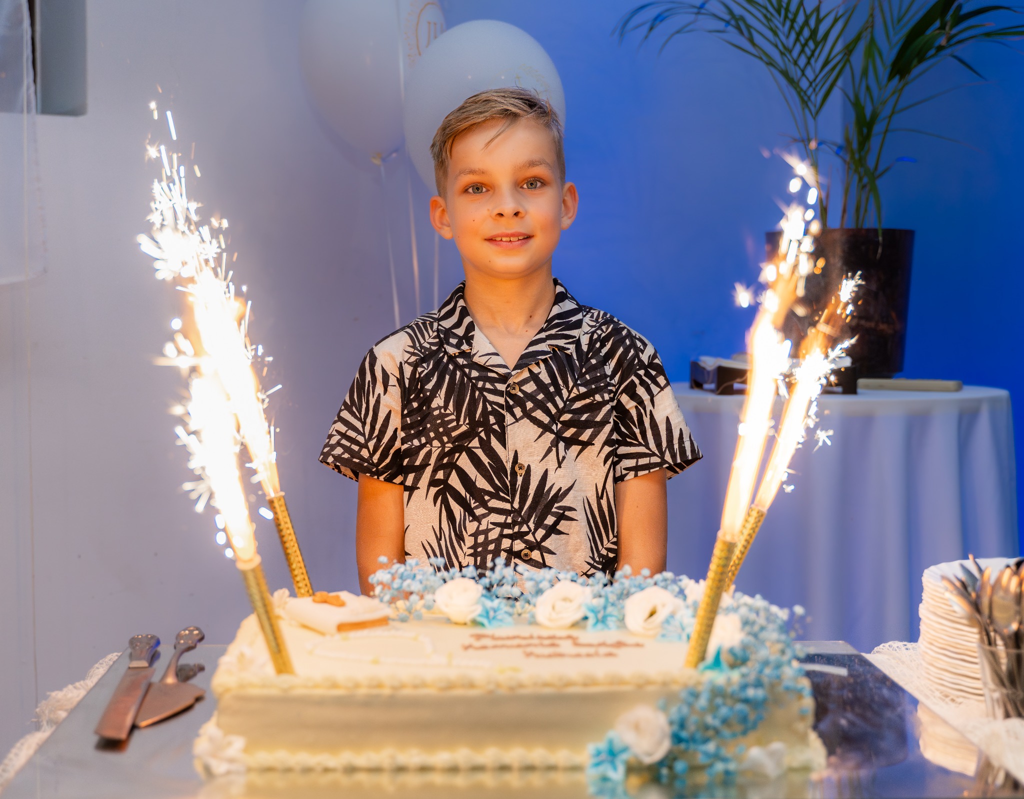 Uśmiechnięty chłopiec w koszuli w liście, stojący za tortem urodzinowym z fontannami iskier, balony i roślina w tle.