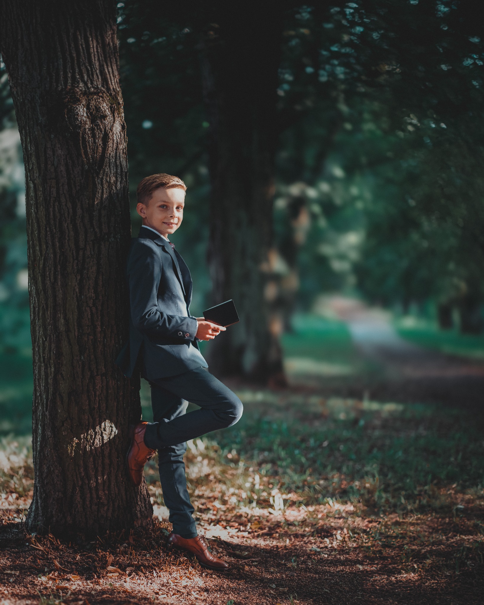 Młody chłopak w granatowym garniturze i brązowych butach opiera się o drzewo w parku, trzymając tablet w dłoni, z uśmiechem patrzy w obiektyw.