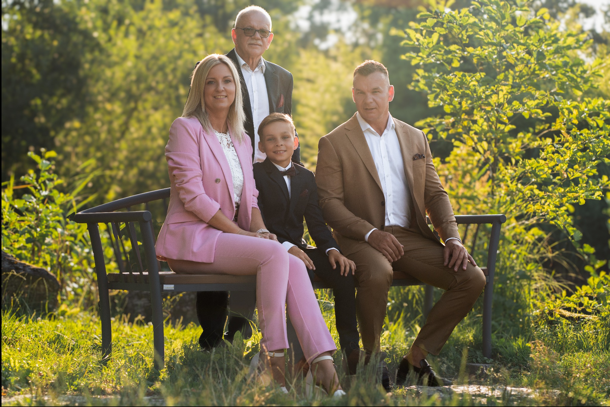 Rodzinne zdjęcie portretowe: kobieta w różowym garniturze, mężczyzna w brązowym garniturze, chłopiec w czarnym garniturze z muszką i starszy mężczyzna w garniturze w tle, siedzący na ławce w parku.