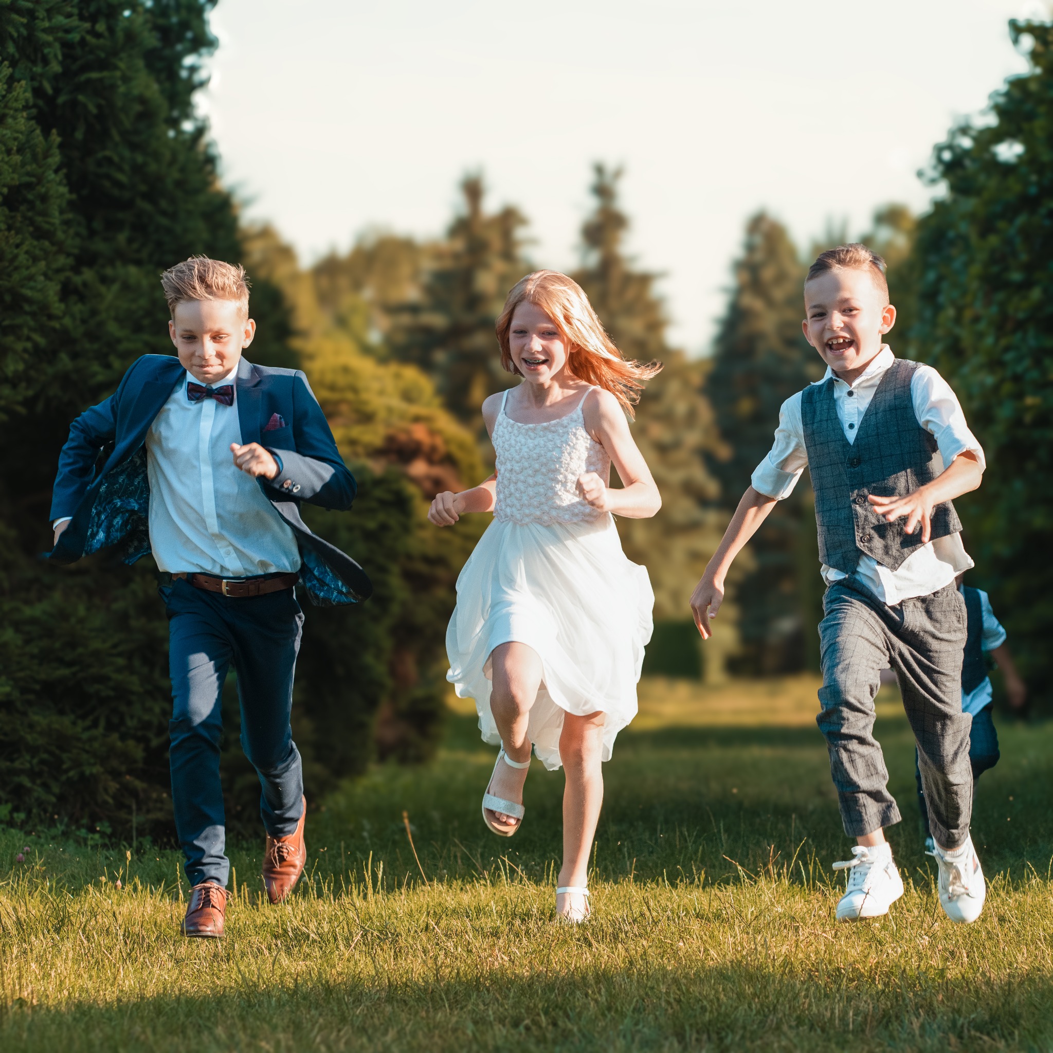 Trójka roześmianych dzieci biegnie po zielonej trawie w parku, ubrana w eleganckie stroje: chłopak w granatowym garniturze z bordową muchą, dziewczynka w białej sukience i chłopak w szarej...