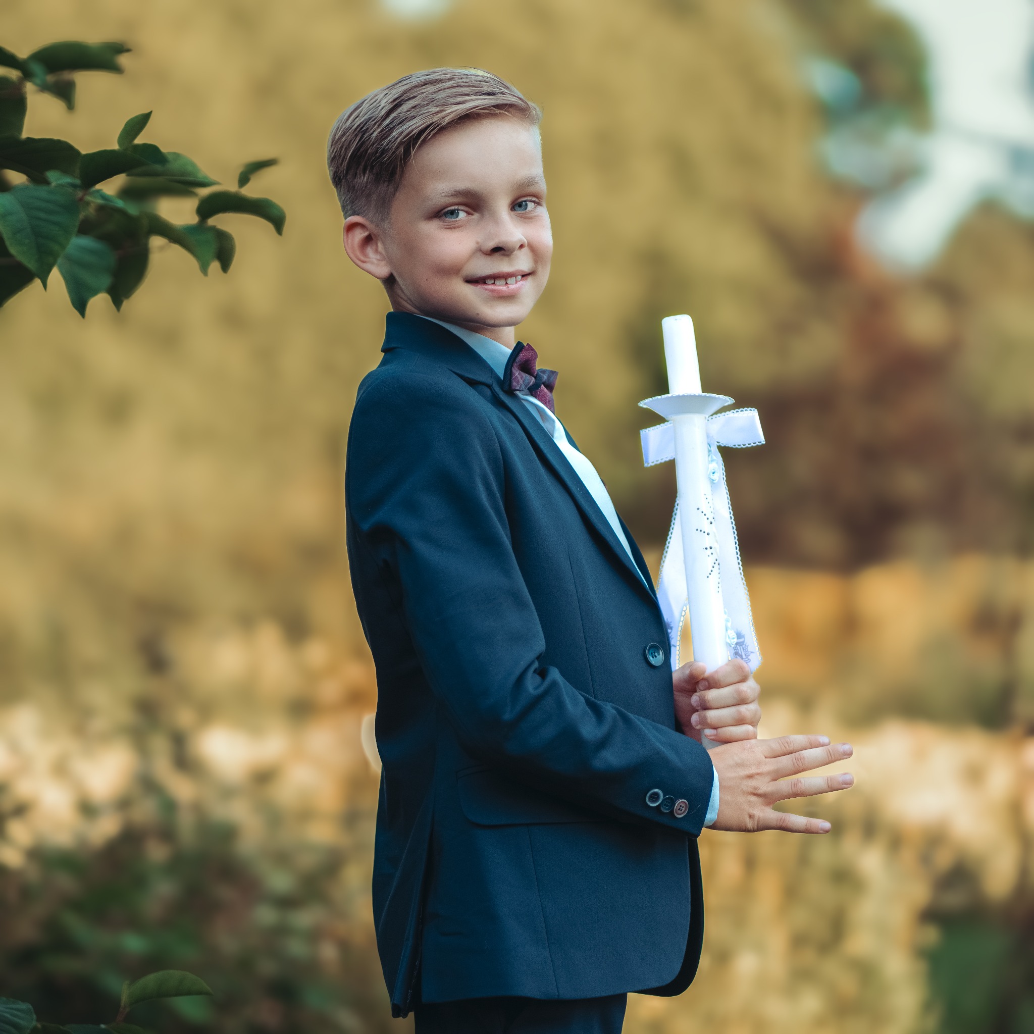Chłopiec w granatowym garniturze i muszce, trzymający świecę komunijną z białymi wstążkami na tle rozmytej zieleni.