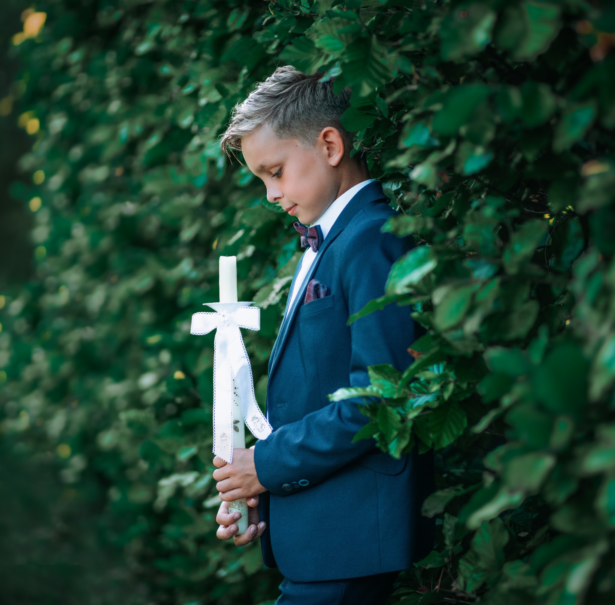 Chłopiec w granatowym garniturze z bordową muchą, trzymający świecę komunijną z białą kokardą na tle zielonych liści.