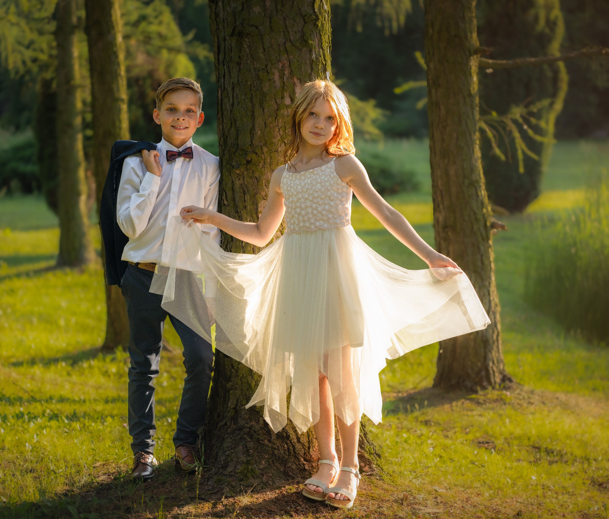 Uśmiechnięty chłopiec w koszuli z muszką i marynarką na ramieniu oraz dziewczynka w białej sukience pozują opierając się o drzewo w słonecznym lesie.
