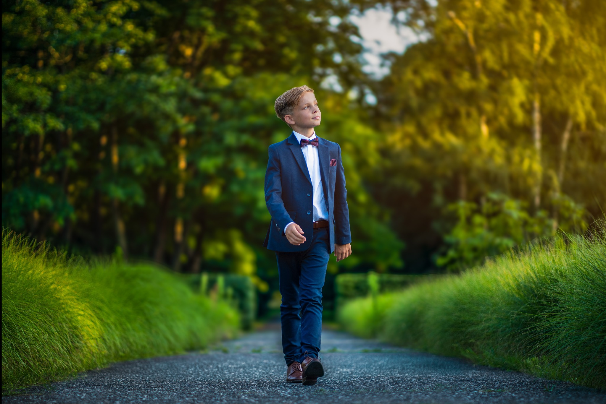 Chłopiec w granatowym garniturze i bordowej muszce idzie ścieżką w parku, spoglądając w górę na słońce.