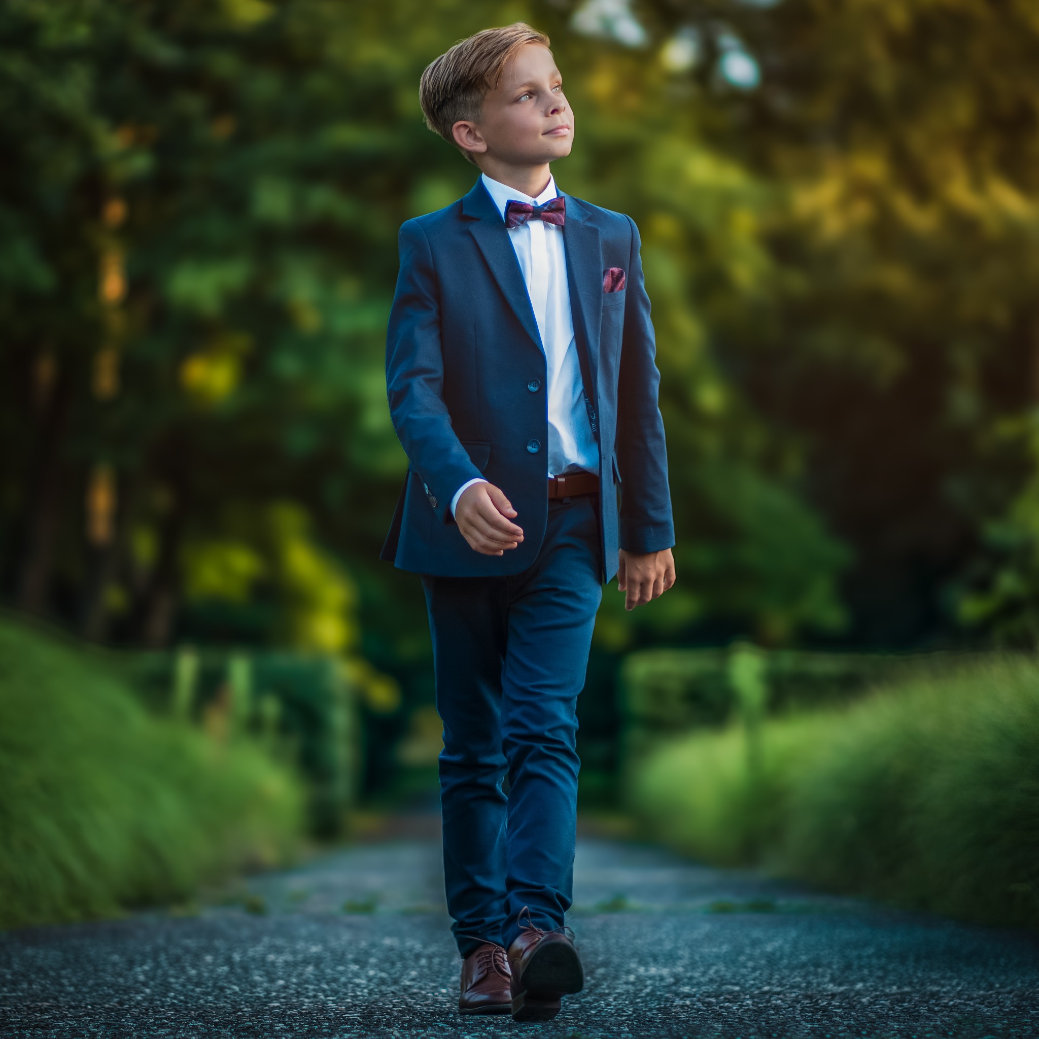 Chłopiec w granatowym garniturze z bordową muchą i poszetką spaceruje leśną ścieżką, spoglądając w górę.