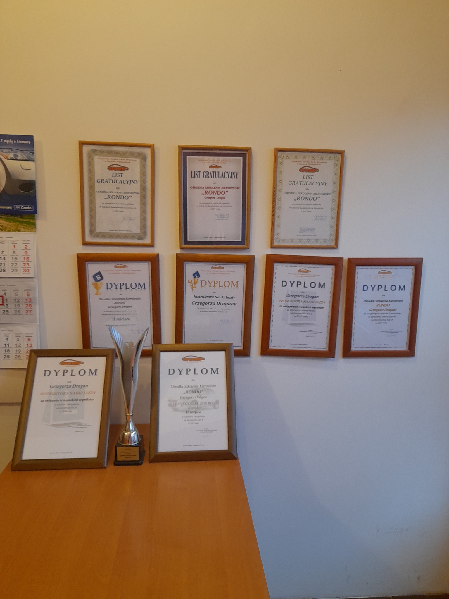 Ściana z ramkami zawierającymi dyplomy i listy gratulacyjne ośrodka szkolenia kierowców 'Rondo', dodatkowo na komodzie stoi srebrna statuetka.