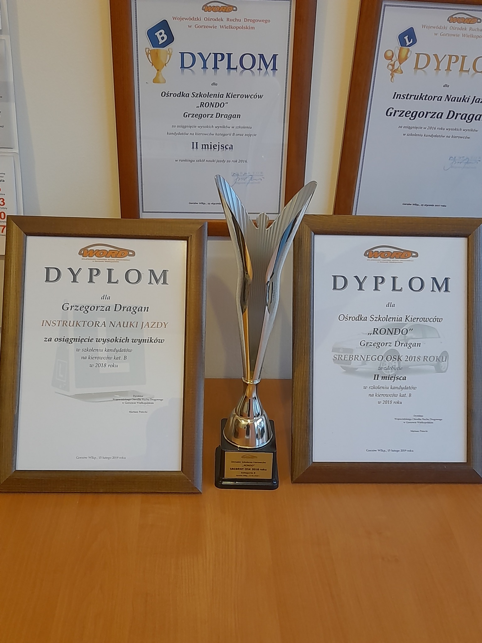Dyplomy i nagroda dla szkoły jazdy 'Rondo' i instruktora Grzegorza Dragana za wysokie wyniki w szkoleniu kierowców, widoczne certyfikaty z lat 2016 i 2018.