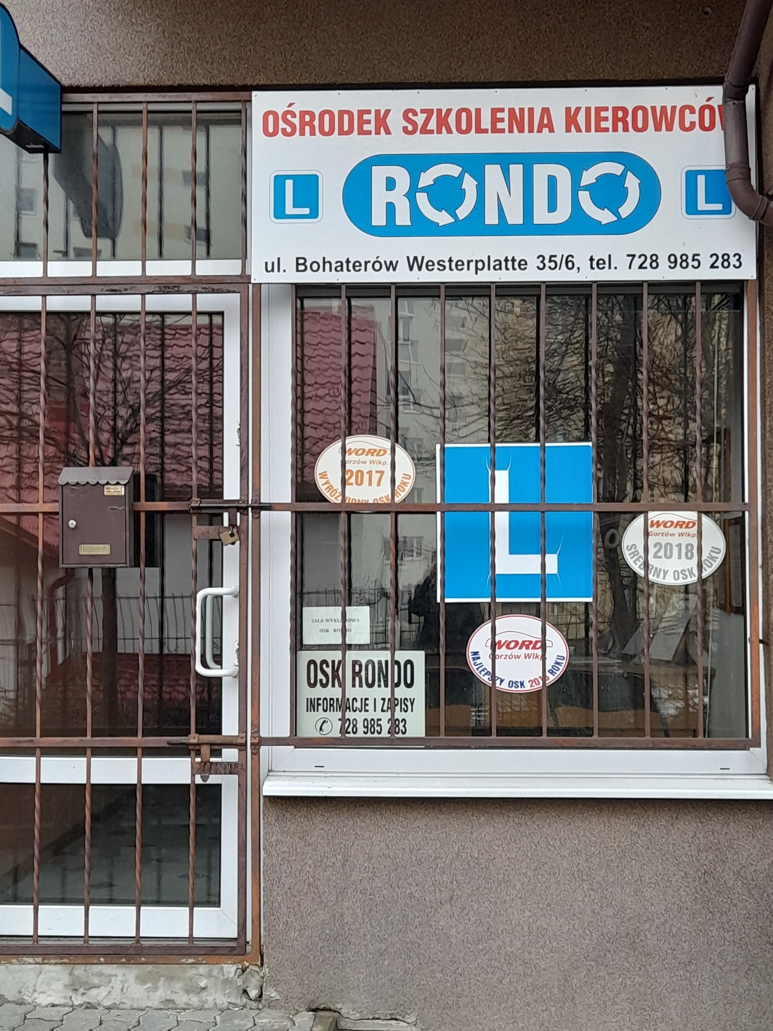 Wejście do Ośrodka Szkolenia Kierowców Rondo z okratowanymi drzwiami i oknem, na którym widnieje szyld z nazwą, adresem i numerem telefonu, a także naklejki z wyróżnieniami z lat 2017, 2018 i 2016.