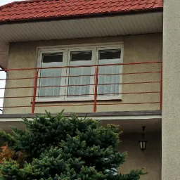 Balkon z pomarańczową, metalową balustradą, widok z dołu na część budynku z oknami i zasłonami, fragment iglastego drzewa zasłaniającego część balkonu.