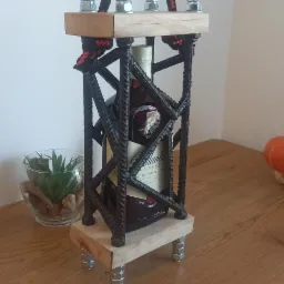 Dekoracyjny stojak na butelkę wykonany z prętów zbrojeniowych, drewnianych elementów i nakrętek, stojący na drewnianym blacie.