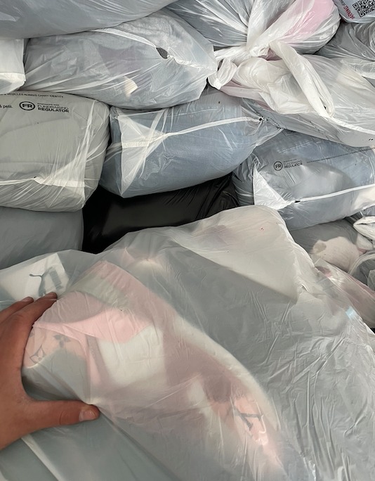 Stos worków foliowych wypełnionych odzieżą, widoczne etykiety i fragmenty ubrań w różnych kolorach.