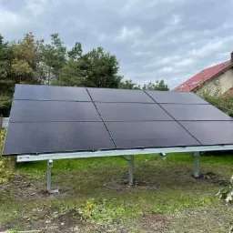 Naziemna instalacja z czarnych paneli słonecznych na metalowym stelażu, widoczna w ogrodzie z budynkiem w tle, częściowo ocieniona przez drzewa.