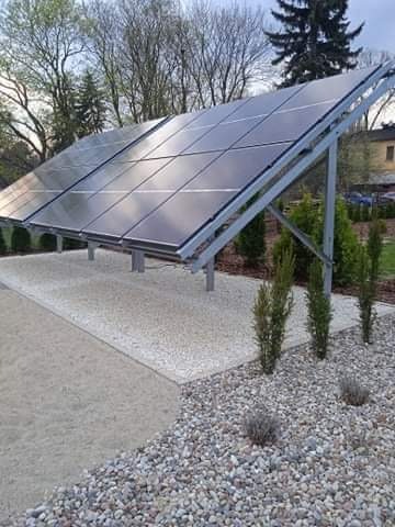 Instalacja paneli słonecznych na metalowej konstrukcji, otoczona dekoracyjnym żwirem i zielenią, na tle drzew i budynku.