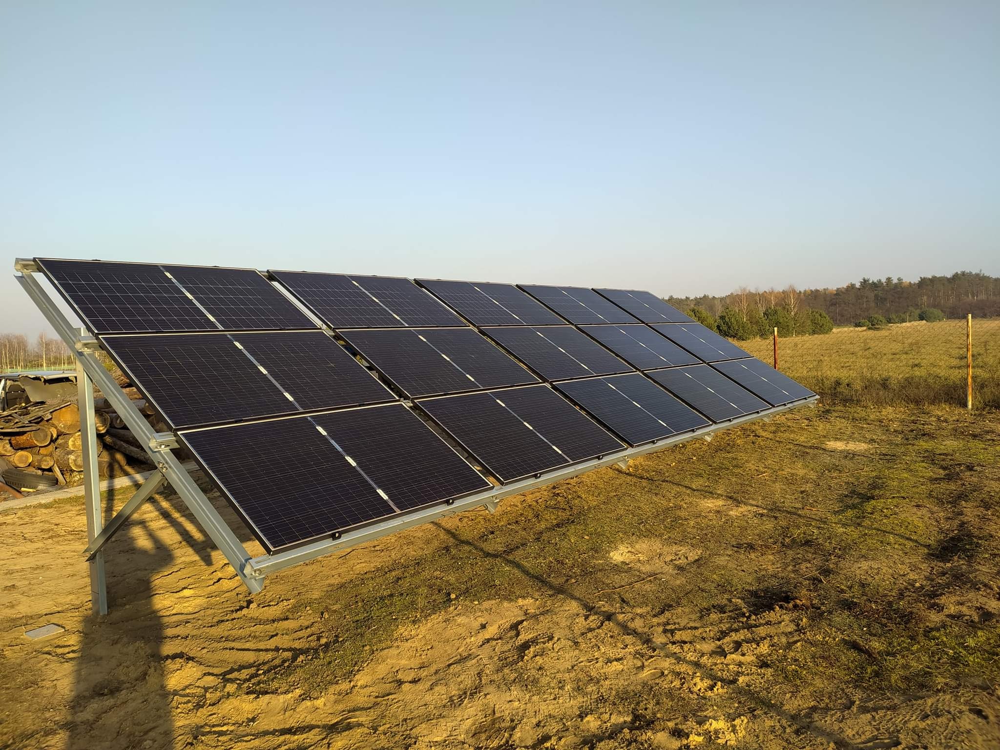 Rząd paneli słonecznych na metalowej konstrukcji, ustawiony na nierównym, piaszczystym terenie z widocznymi śladami opon i stertą drewna w tle, pod bezchmurnym niebem.