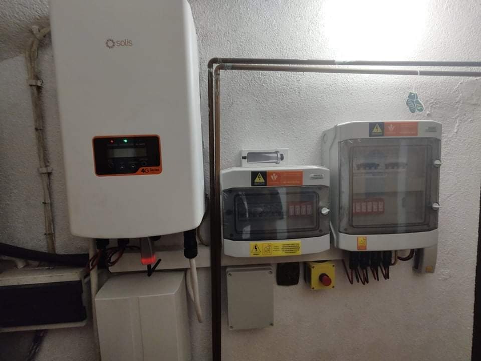 Biały falownik Solis zamontowany na ścianie obok dwóch szarych skrzynek bezpiecznikowych z przewodami i żółtym przyciskiem awaryjnym.