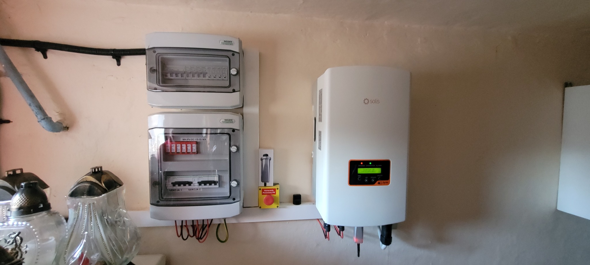 Zamontowany biały falownik Solis 4G Series z zielonym wyświetlaczem, obok dwie skrzynki bezpiecznikowe Noark MH-PV-DC-1S-1S-MX i wyłącznik pożarowy PV na beżowej ścianie wewnątrz budynku.