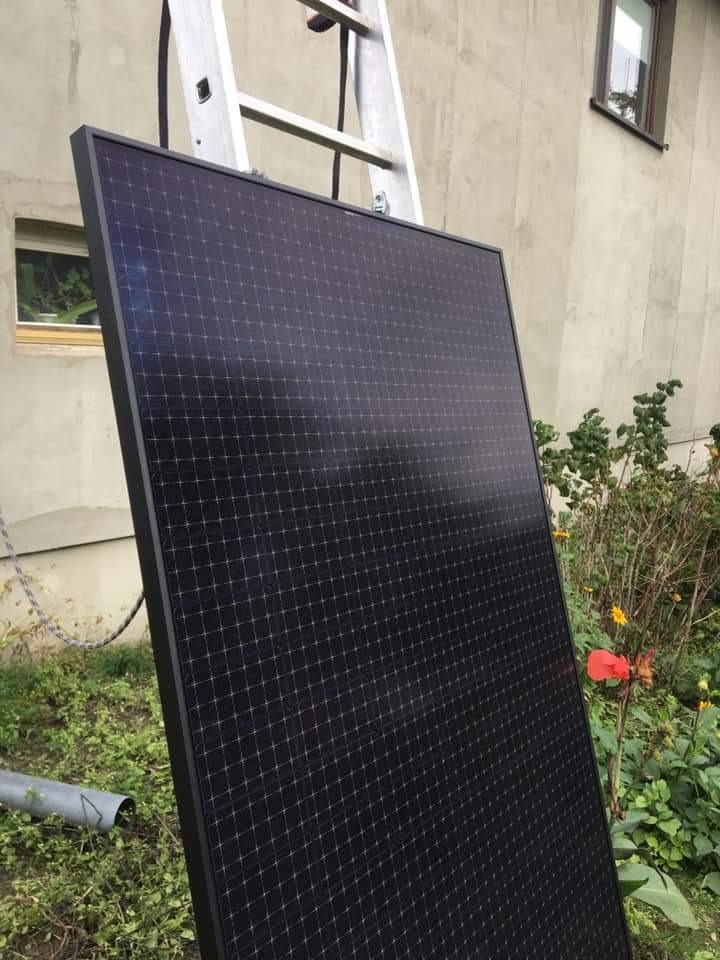 Panel słoneczny oparty o drabinę na tle ściany budynku i zieleni, widoczny fragment okna.