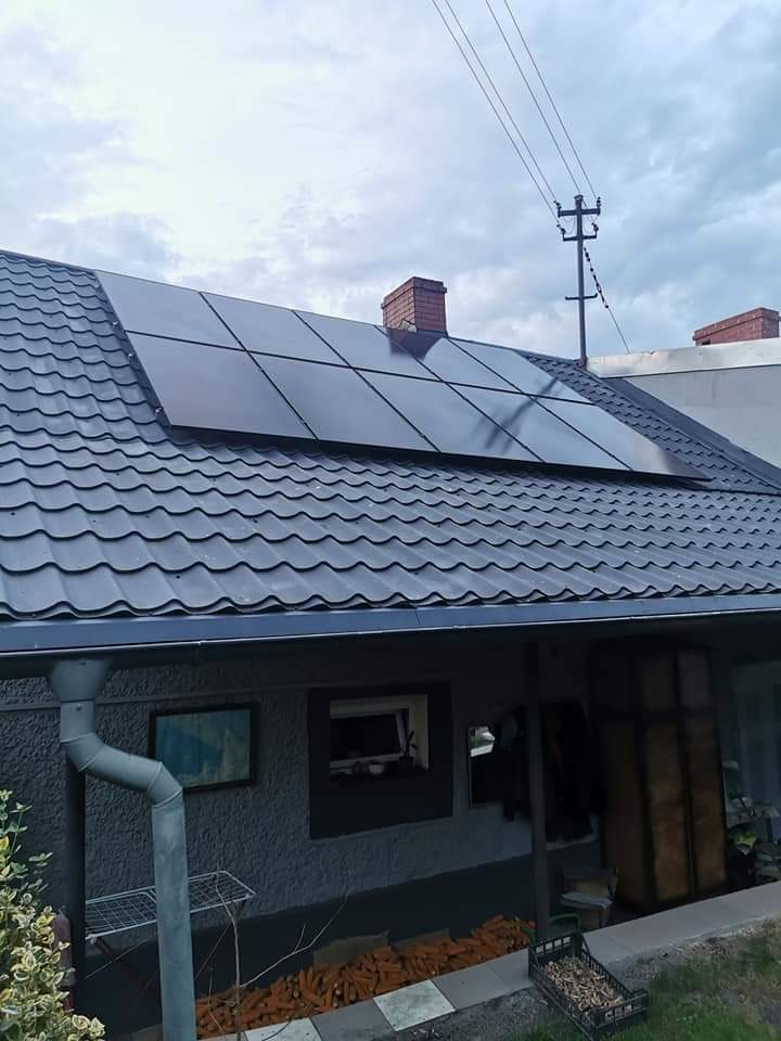 Czarny panel słoneczny na ciemnym, metalowym dachu domu jednorodzinnego w pochmurny dzień, z widoczną rynną i stertą kukurydzy pod okapem.