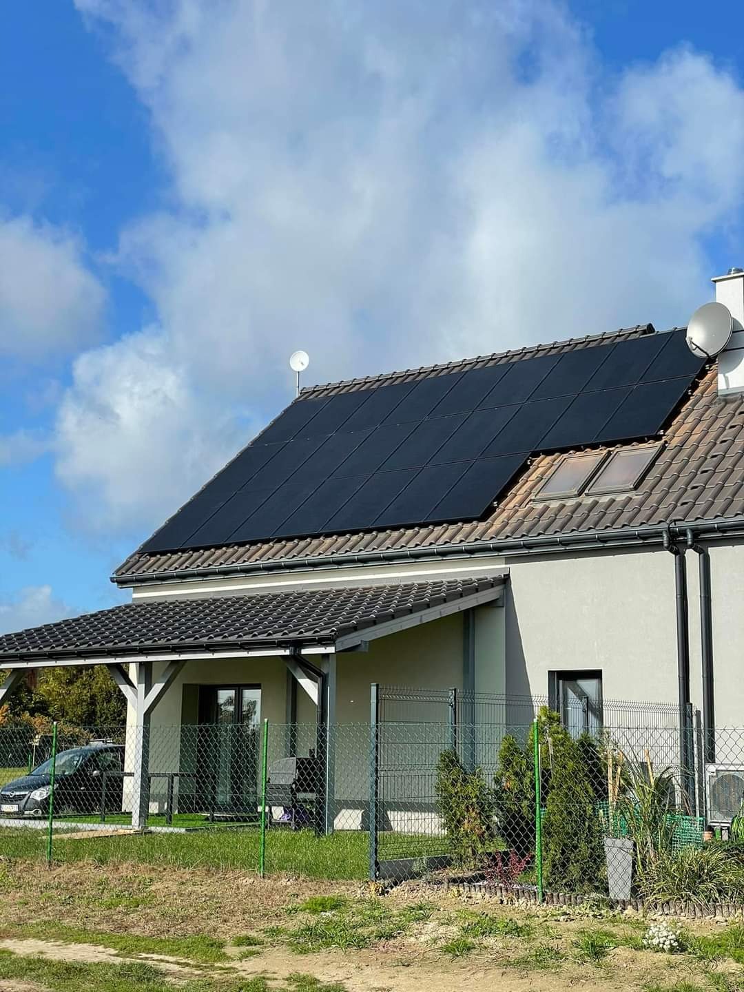 Dom jednorodzinny z panelami słonecznymi na dachu, widoczne anteny satelitarne oraz ogród z zielonym ogrodzeniem.