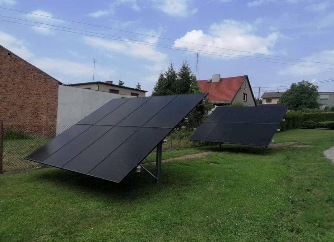 Dwa panele słoneczne ustawione na trawniku przed domem jednorodzinnym, widoczne elementy konstrukcyjne mocujące.