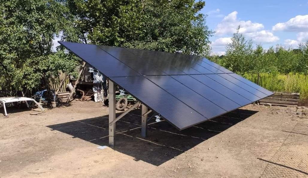 Czarny panel słoneczny zamontowany na metalowej konstrukcji, na tle wiejskiego krajobrazu z widocznymi drzewami i złomem.