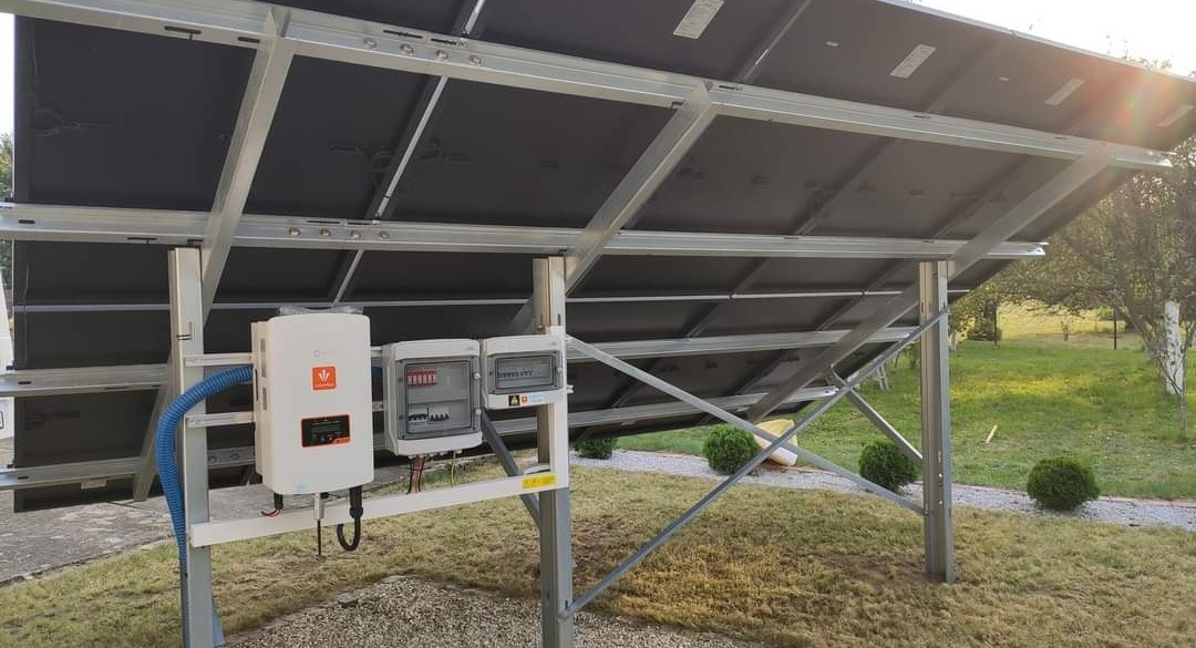 Widok od spodu na panele słoneczne zamontowane na metalowej konstrukcji, z widocznym inwerterem i skrzynką przyłączeniową, na tle trawnika z krzewami.