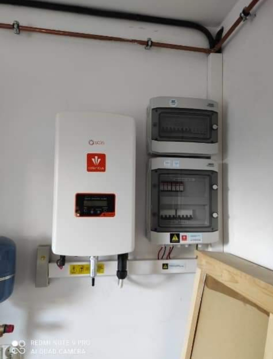 Biały falownik Solis zamontowany na ścianie obok dwóch szarych rozdzielnic z widocznymi bezpiecznikami, instalacja w pomieszczeniu technicznym z rurami miedzianymi pod sufitem.