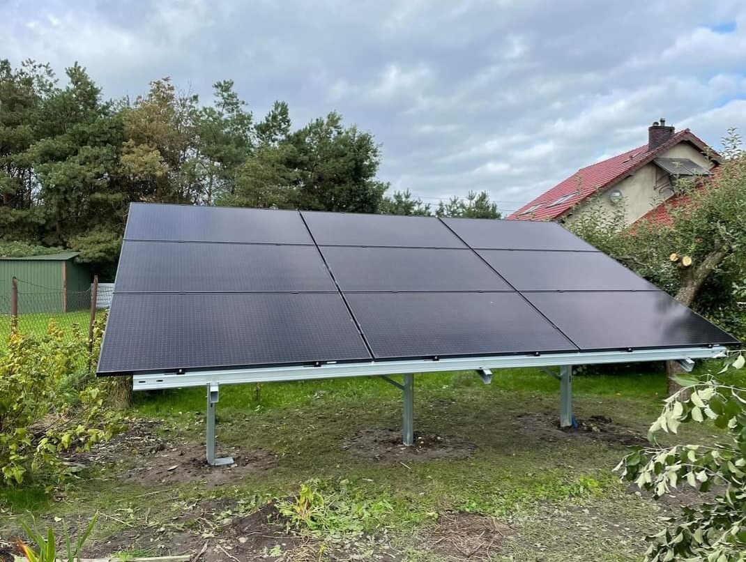 Naziemna instalacja z czarnych paneli słonecznych na metalowym stelażu, widoczna w ogrodzie z budynkiem w tle, częściowo ocieniona przez drzewa.