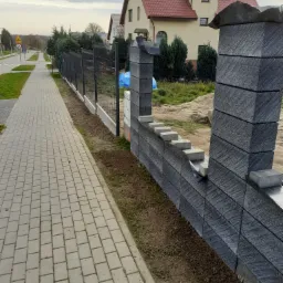 Nowoczesne ogrodzenie z grafitowych bloczków betonowych w trakcie montażu, widoczna metalowa siatka i podmurówka, tło stanowi chodnik z kostki i dom z czerwoną dachówką.