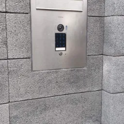 Zewnętrzny panel domofonowy Vidos z klawiaturą numeryczną i kamerą, zamontowany na szarej ścianie z bloczków betonowych.