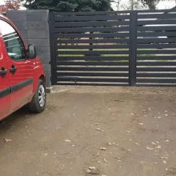 Nowoczesna, pozioma brama wjazdowa z szarych betonowych bloczków, widziana z boku czerwonego samochodu dostawczego z naklejkami informującymi o usługach: Matyka Bram, Monitoring, Domofony, Alarmy...