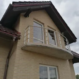 Kremowy, dwupiętrowy dom z cegły klinkierowej z balkonem zabezpieczonym metalową, półokrągłą barierką, widok z dołu.