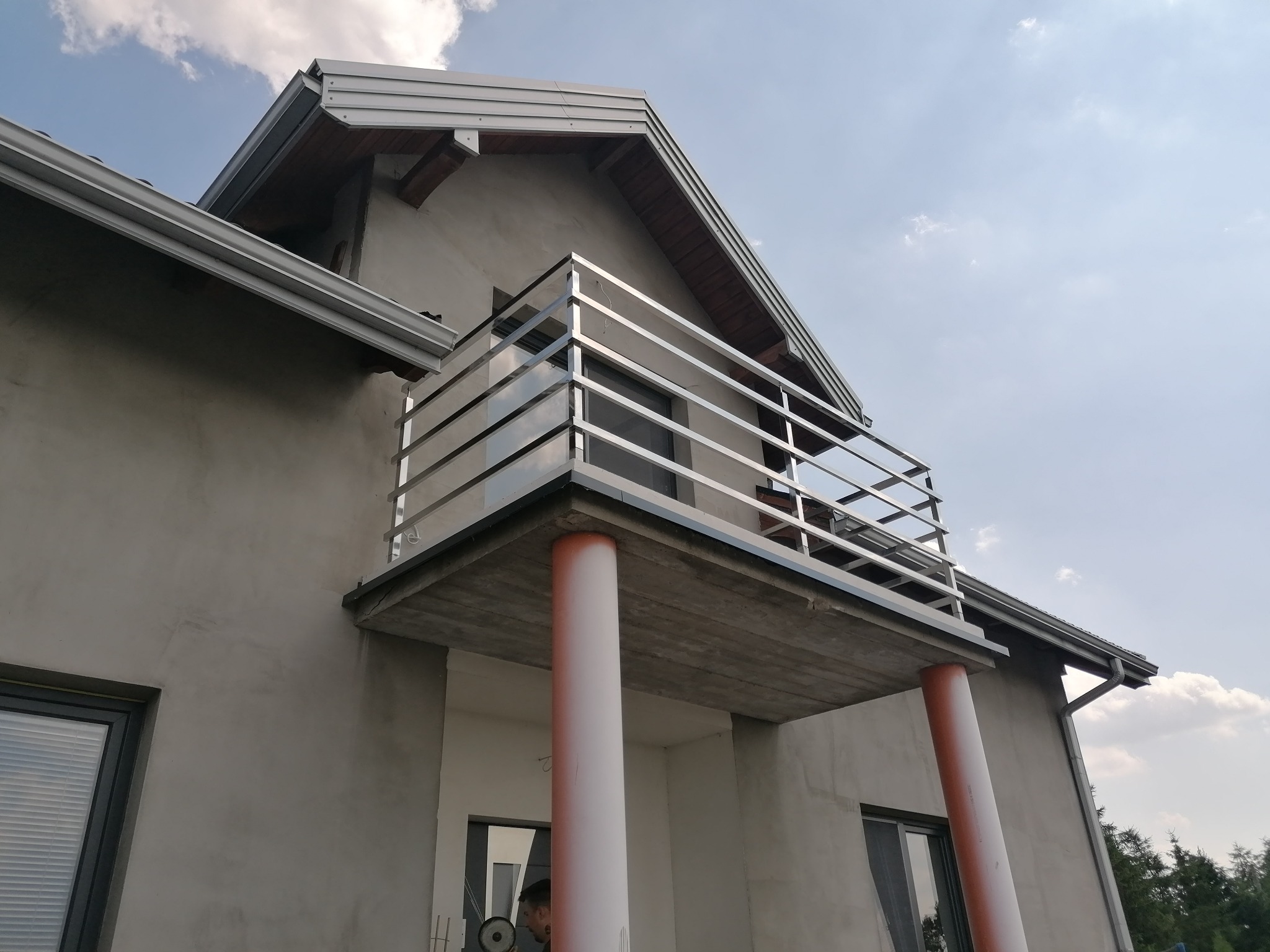 Balkon z poziomą, metalową balustradą, wsparty na dwóch kolumnach, widok z dołu na tle nieba i częściowo otynkowanej ściany budynku.