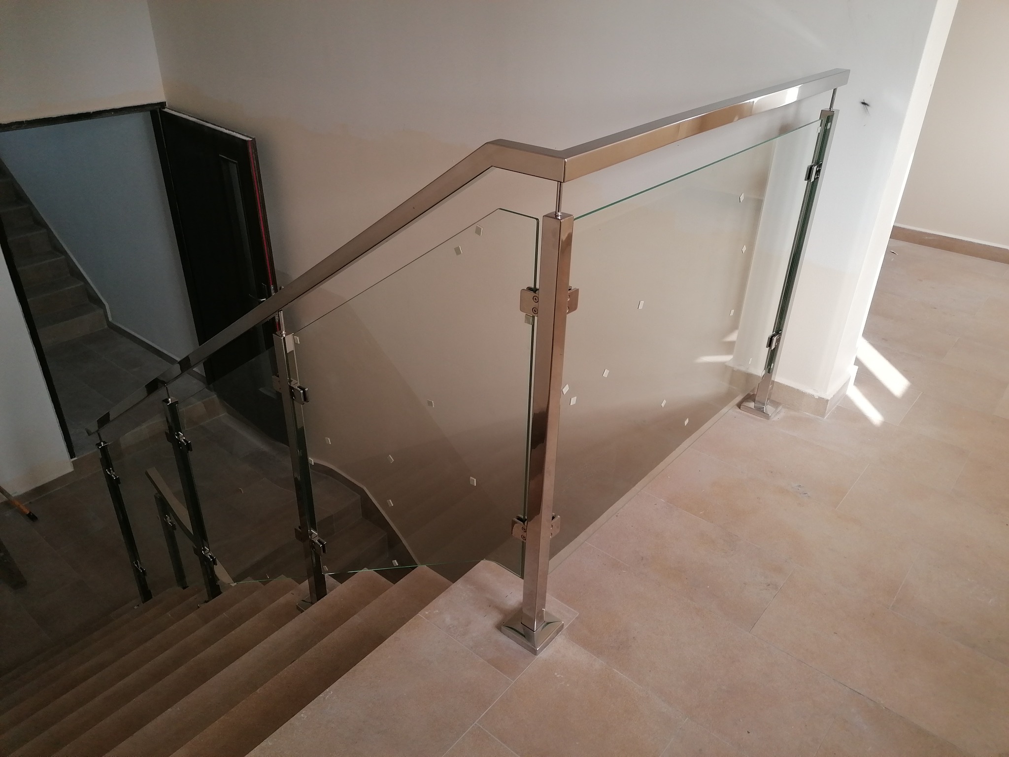 Szklana balustrada ze stali nierdzewnej na schodach z beżowych płytek, widok z góry, w tle widoczne drzwi.