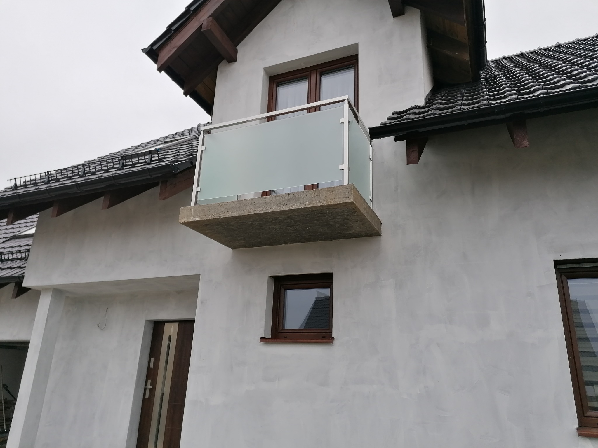 Balkon z balustradą ze szkła matowego i stalową ramą, zamontowany na betonowej płycie, widok z dołu na elewację domu z ciemnobrązowymi oknami i dachem z dachówki ceramicznej.
