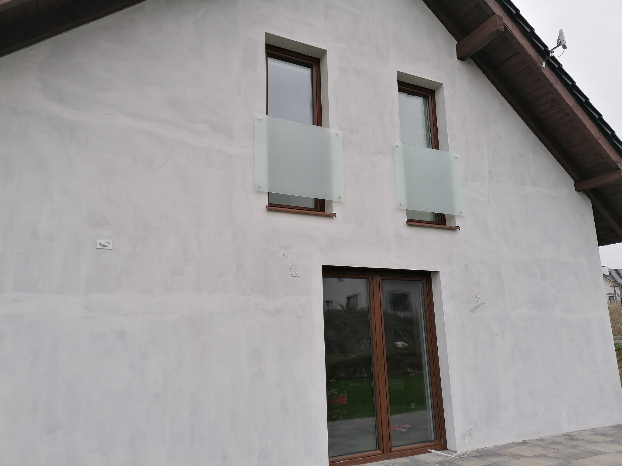 Elewacja domu z oknami zabezpieczonymi panelami z matowego szkła, widoczne elementy drewniane dachu i szara kostka brukowa na podjeździe.