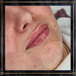 Zbliżenie na usta po zabiegu pigmentacji, widoczny efekt błyszczących, pełnych ust w odcieniu różu, z logo salonu kosmetycznego w prawym dolnym rogu.