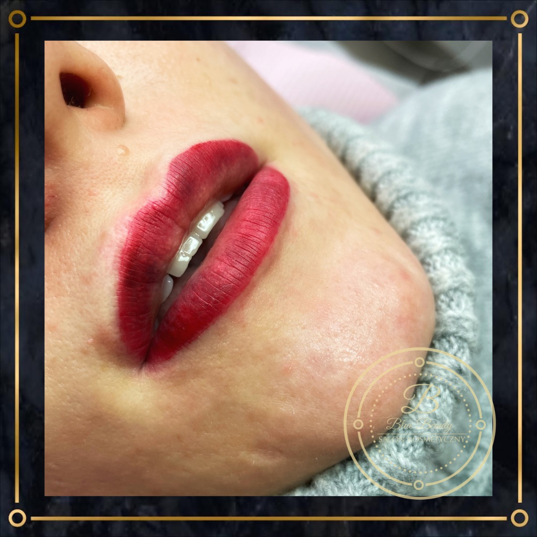 Zbliżenie na usta po zabiegu makijażu permanentnego, z wyraźnie zarysowanym konturem i intensywnym czerwonym kolorem, widoczny fragment twarzy i szary sweter, w rogu logo salonu kosmetycznego.