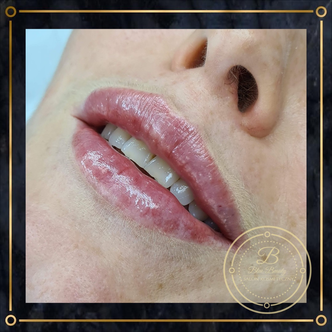 Zbliżenie na usta po zabiegu pigmentacji, widoczne świeże, błyszczące usta w odcieniu różu i fragment zębów, w prawym dolnym rogu logo salonu kosmetycznego.