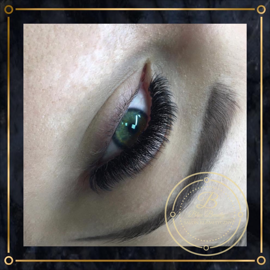 Zbliżenie na oko z gęstymi, czarnymi, profesjonalnie przedłużonymi rzęsami; widoczny fragment brwi i logo salonu kosmetycznego w rogu.