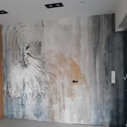 Ściana z fototapetą przedstawiającą baletnicę w zwiewnej sukni na tle abstrakcyjnych, kolistych wzorów w odcieniach szarości, beżu i błękitu, z widocznymi elementami instalacji elektrycznej.