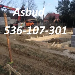 Fundamenty domu w trakcie budowy: szalunki drewniane, palety z bloczkami betonowymi, dźwig Palfinger i pomarańczowy bus w tle.