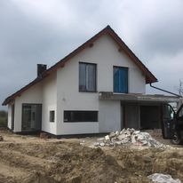 Surowy stan dwukondygnacyjnego domu jednorodzinnego z białymi ścianami zewnętrznymi i ciemnymi oknami, widoczne ślady prac budowlanych wokół budynku, częściowo zadaszone wejście, pochmurne niebo...