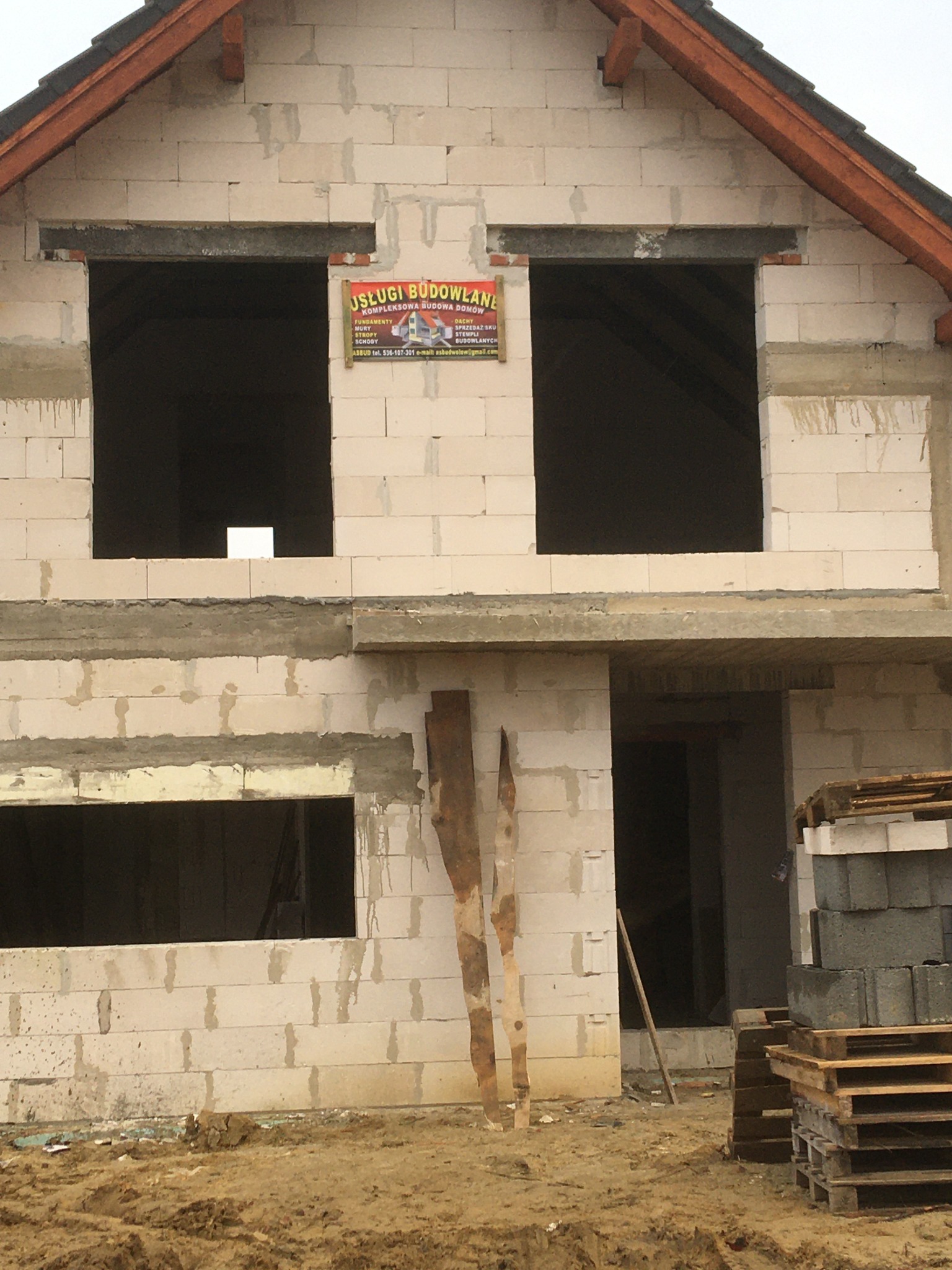 Niedokończony budynek z betonu komórkowego z widocznymi otworami okiennymi i drzwiowymi, na ścianie wisi tablica reklamowa usług budowlanych, na pierwszym planie ziemia i palety z bloczkami.