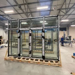 Gotowe moduły okienne z czarnymi ramami, ustawione na paletach w hali produkcyjnej, widoczne napisy 'ALUPROF' na profilach, w tle pracownik przy stanowisku.