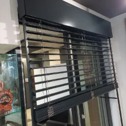 Ciemne żaluzje fasadowe zamontowane w oknie z czarną ramą, widoczne sznurki sterujące i fragment ekspozycji sklepowej z gablotą.