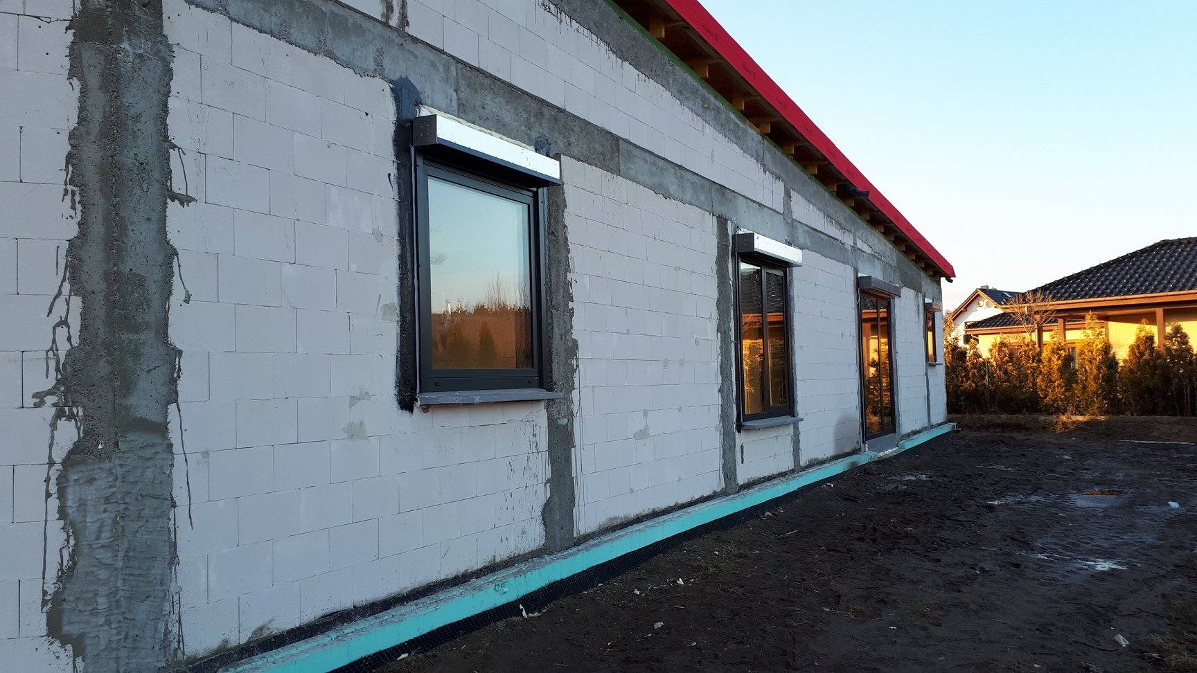 Surowy budynek z szarego betonu komórkowego w trakcie budowy, z zamontowanymi oknami z roletami zewnętrznymi, widoczne ślady zaprawy murarskiej i izolacja przeciwwilgociowa przy gruncie.